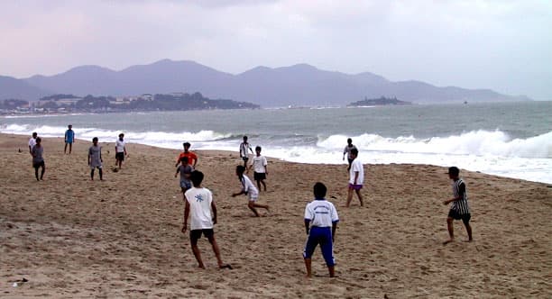 People playing soccer on Nha Trang Beach with clear turquoise water and coastal resort developments visible in the background.