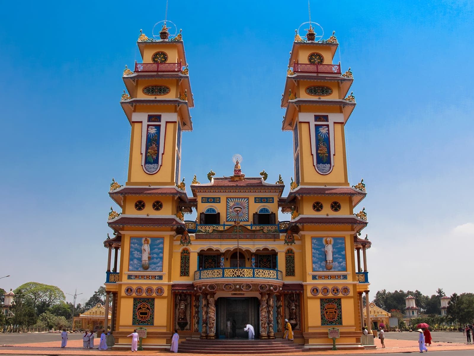 The Cao Dai Holy See temple displaying distinctive pink, blue, and yellow striped exterior with ornate architectural details and pointed spires against a clear sky.