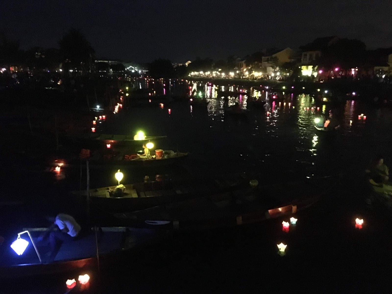 Illuminated paper lanterns floating on water in Hoi An's ancient town during the full moon festival night, with colorful reflections shimmering on the riverbank
