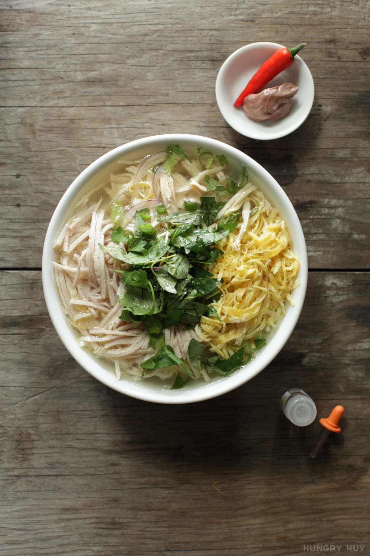 A clear broth noodle soup in a white bowl, garnished with shredded egg, chicken, and ham ribbons.