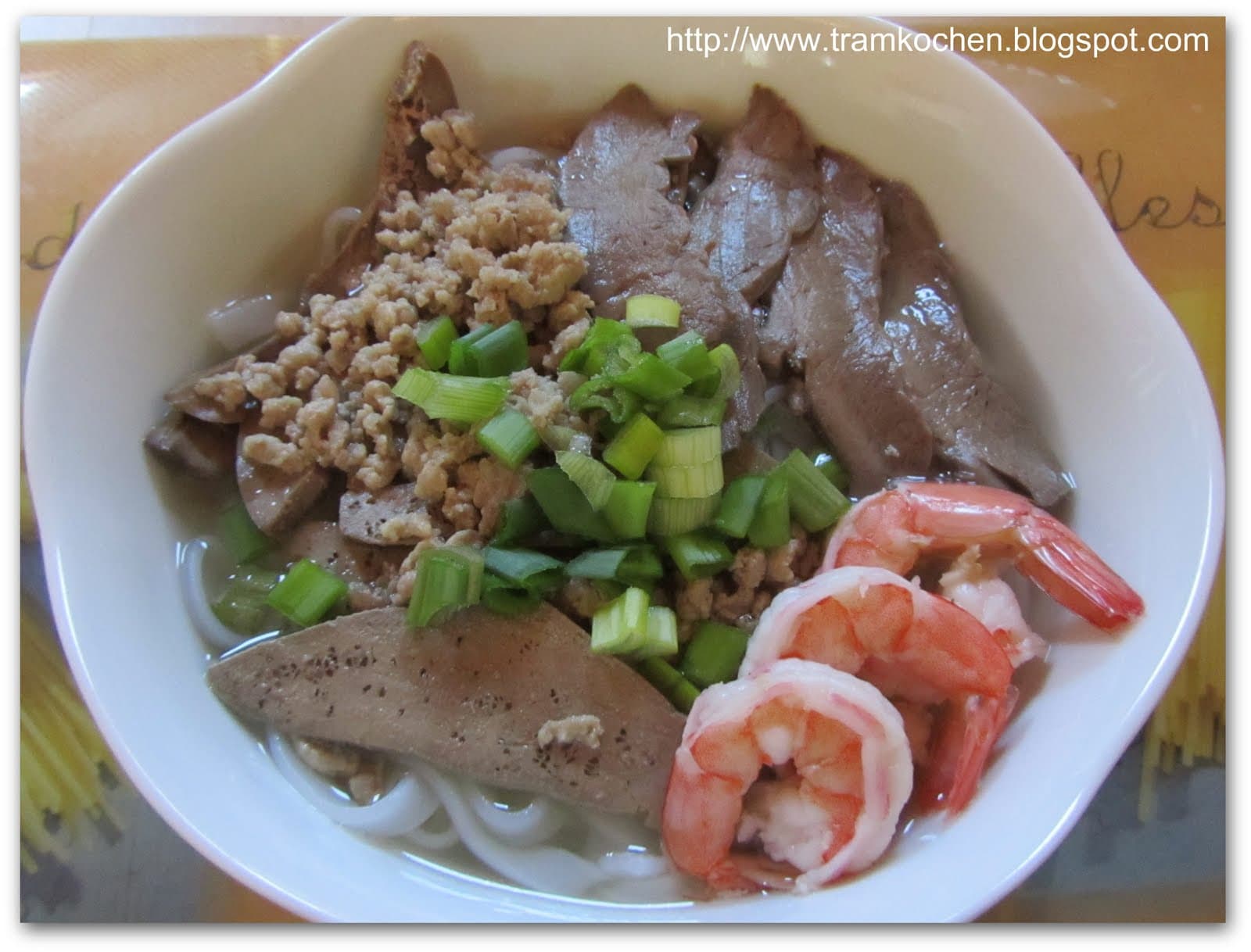 A steaming bowl of clear pork broth noodle soup with prawns, pork, crackling, and fresh herbs on a dark table