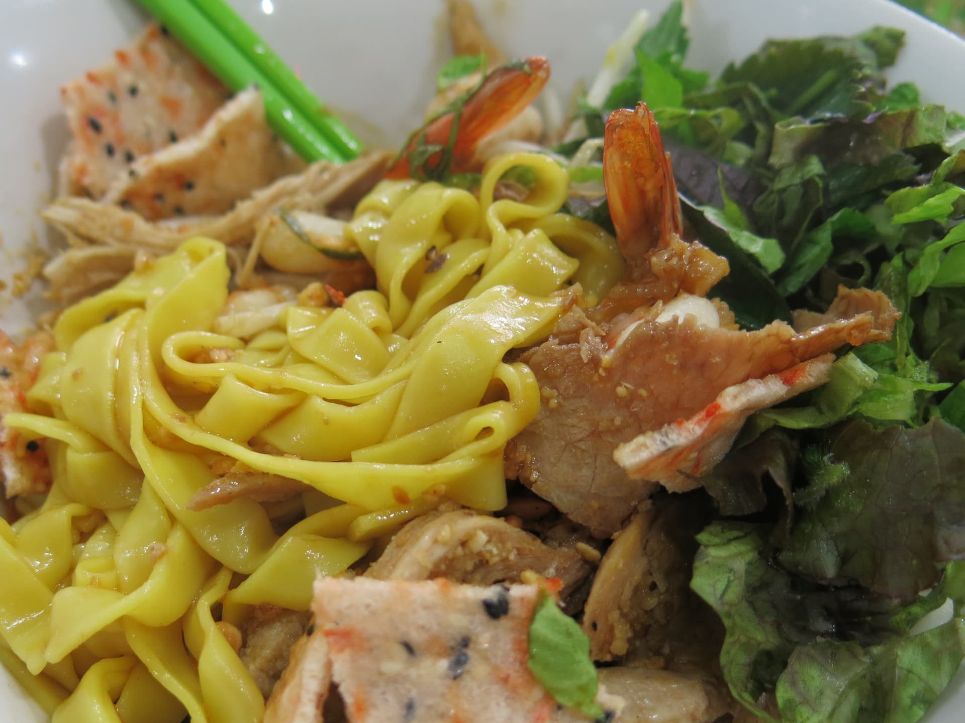 Bright yellow turmeric rice noodles in a shallow bowl with pork, shrimp, peanuts, herbs, and grilled rice cracker.