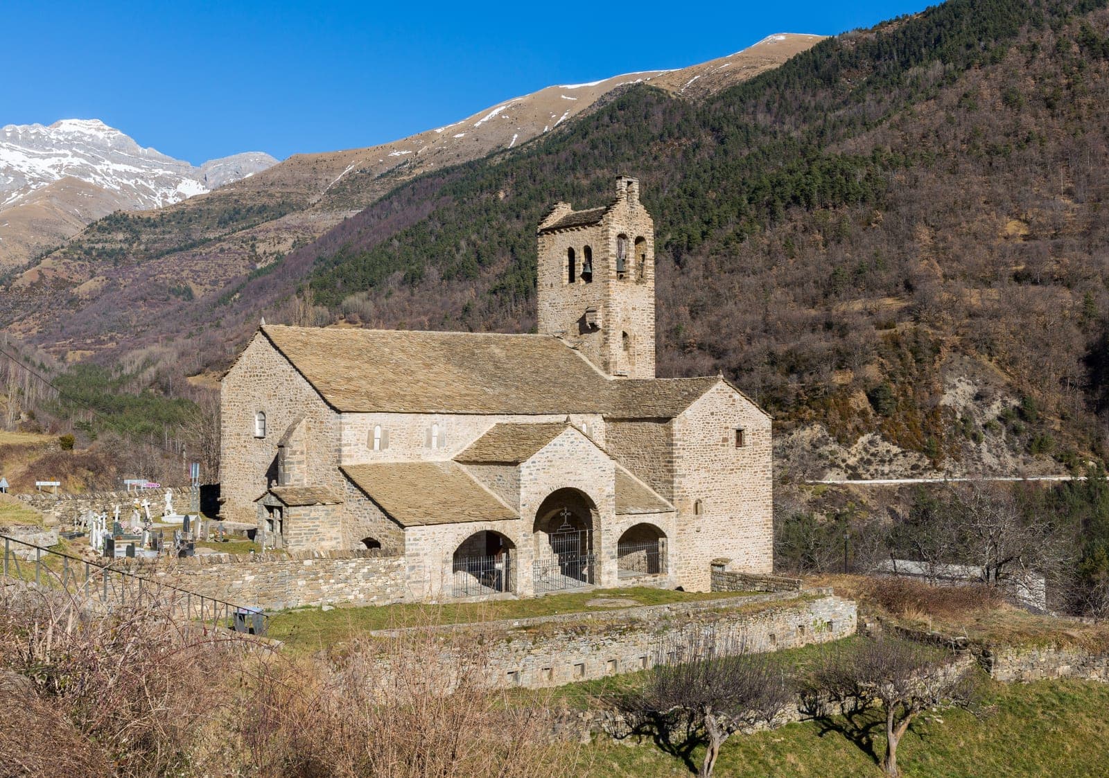 Stone Romanesque church with square bell tower set in a forested mountain valley, surrounded by snow-capped Pyrenean peaks and pine forests